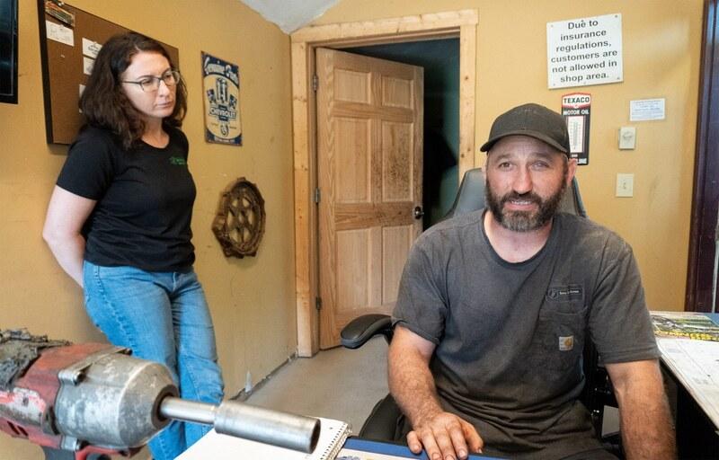 Ross Tobiassen sits in a chair inside a home office. His wife Rebecca looks at him.